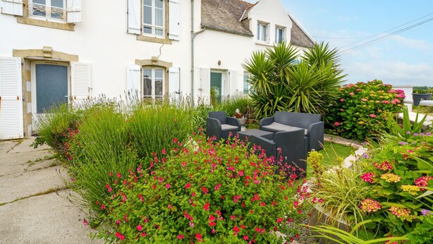 A white house with shutters, a garden with various plants and flowers, and outdoor furniture.