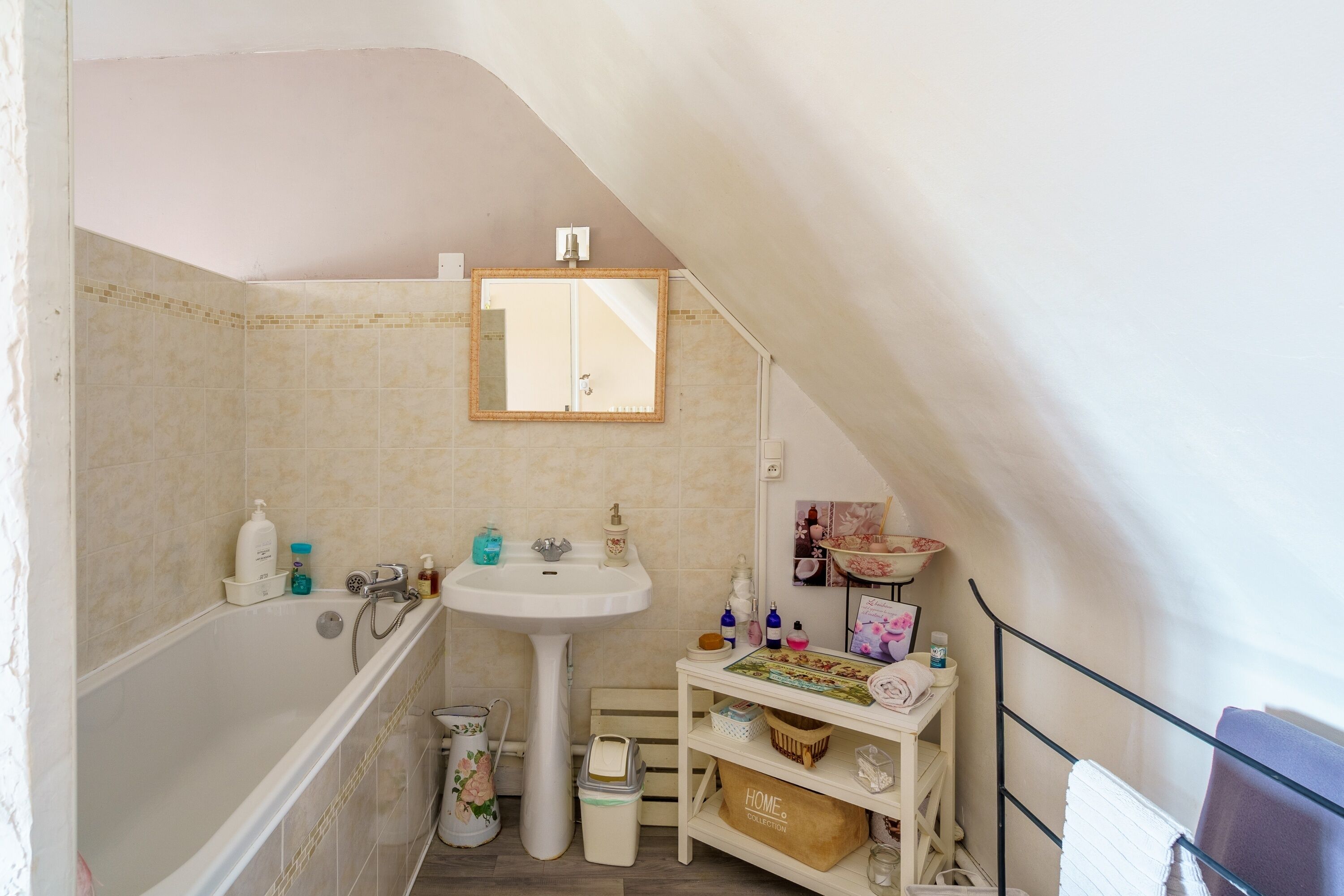 Ein kleines, gemütliches Badezimmer mit Badewanne, Waschbecken und Spiegel. Auf dem Waschbecken stehen Pflegeartikel und ein Regal mit verschiedenen Gegenständen.