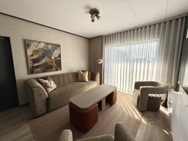Una sala de estar moderna con un sofá, una mesa de centro y dos sillones. Hay un cuadro en la pared y una lámpara en el techo.