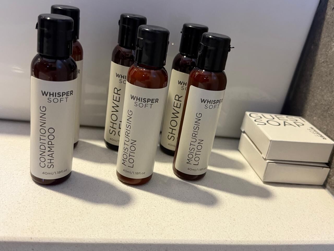 Cinco frascos de produtos Whisper Soft, incluindo condicionador e loção para o banho, e uma caixa de sabonete para hóspedes.
