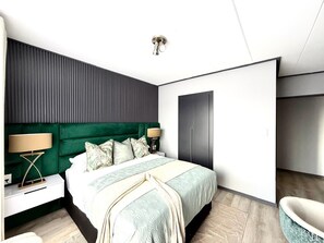 Un dormitorio moderno con un cabecero tapizado en verde, una cama grande con colcha blanca y motivos verdes, dos lámparas de noche y un armario oscuro.