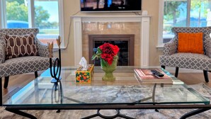 Un salon avec deux canapés à motifs, une table basse en verre, un vase avec des fleurs, une boîte de mouchoirs et une télécommande. Un téléviseur est fixé au-dessus d’une cheminée.
