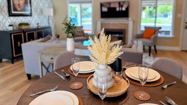 Un coin repas avec une table ronde dressée pour quatre personnes, un vase contenant des plantes séchées et un téléviseur en arrière-plan.