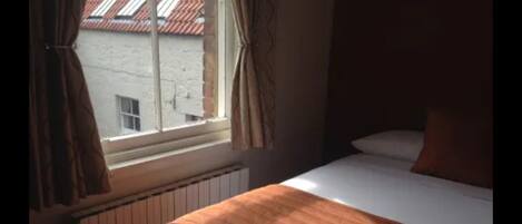 A room with a bed, a window with curtains, and a view of a building with a red roof.