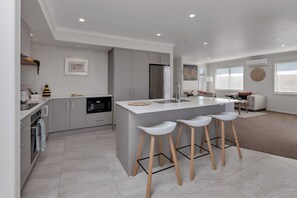 A modern kitchen with a central island, bar stools, and a dining area with a sofa and table. - Serendipity - One Tree Point Holiday Home (Whangarei)