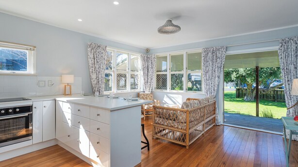 Een keuken met een centraal eiland, witte kasten, een ingebouwde oven en een eethoek met een rieten stoel en tafel.