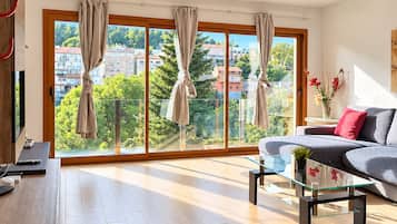Una sala de estar moderna con un sofá, una mesa de centro de vidrio y vistas a un paisaje urbano a través de amplios ventanales.