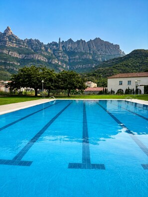 A swimming pool with clear blue water, surrounded by a grassy area, trees, and a building with a red roof. In the background, there are mountains with rocky cliffs. - Private penthouse with jacuzzi, terrace and Montserrat views (Monistrol de Montserrat)