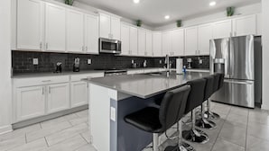 A modern kitchen with white cabinets, a stainless steel refrigerator, a microwave, and a large island with bar stools. - Star Wars & Tree House • Pool & Spa • Game Room • Near Disney (Kissimmee)