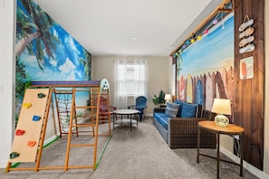 A room with a climbing wall, a sofa, a small table, and a wall with surfboards.