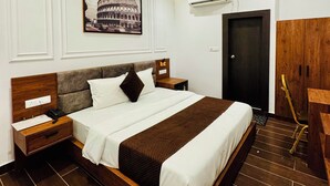 A modern hotel room with a large bed, wooden headboard, a nightstand, a desk, and a chair. - Hotel Ellora Inn Sultanpur (Sultanpur)