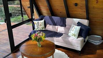Une pièce chaleureuse avec un plafond en bois, un canapé agrémenté de coussins à motifs floraux, une table ronde en bois surmontée d’un chemin de table en crochet, et une fenêtre donnant sur un paysage verdoyant.