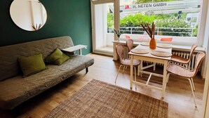 Ett modernt vardagsrum med en grön accentvägg, en rund spegel, en grå soffa med gröna kuddar, en vävd matta och en matplats med flätade stolar och ett bord.