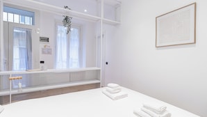 Un baño moderno y limpio con un lavamanos blanco, un espejo y una ventana con cortinas translúcidas. Hay toallas en la encimera y una obra de arte enmarcada en la pared.