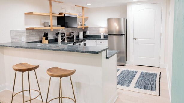 A modern kitchen with a central island, bar stools, a stainless steel refrigerator, and wooden shelves. - 4-Bedroom Home with Fenced Backyard (Lander)