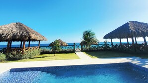 Cielo azul claro, piscina y dos construcciones con techo de paja con vista al océano.