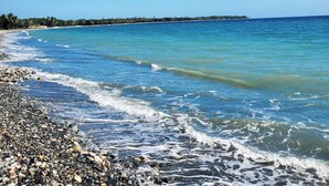 Una playa con agua azul cristalina, piedras y una costa lejana con vegetación.