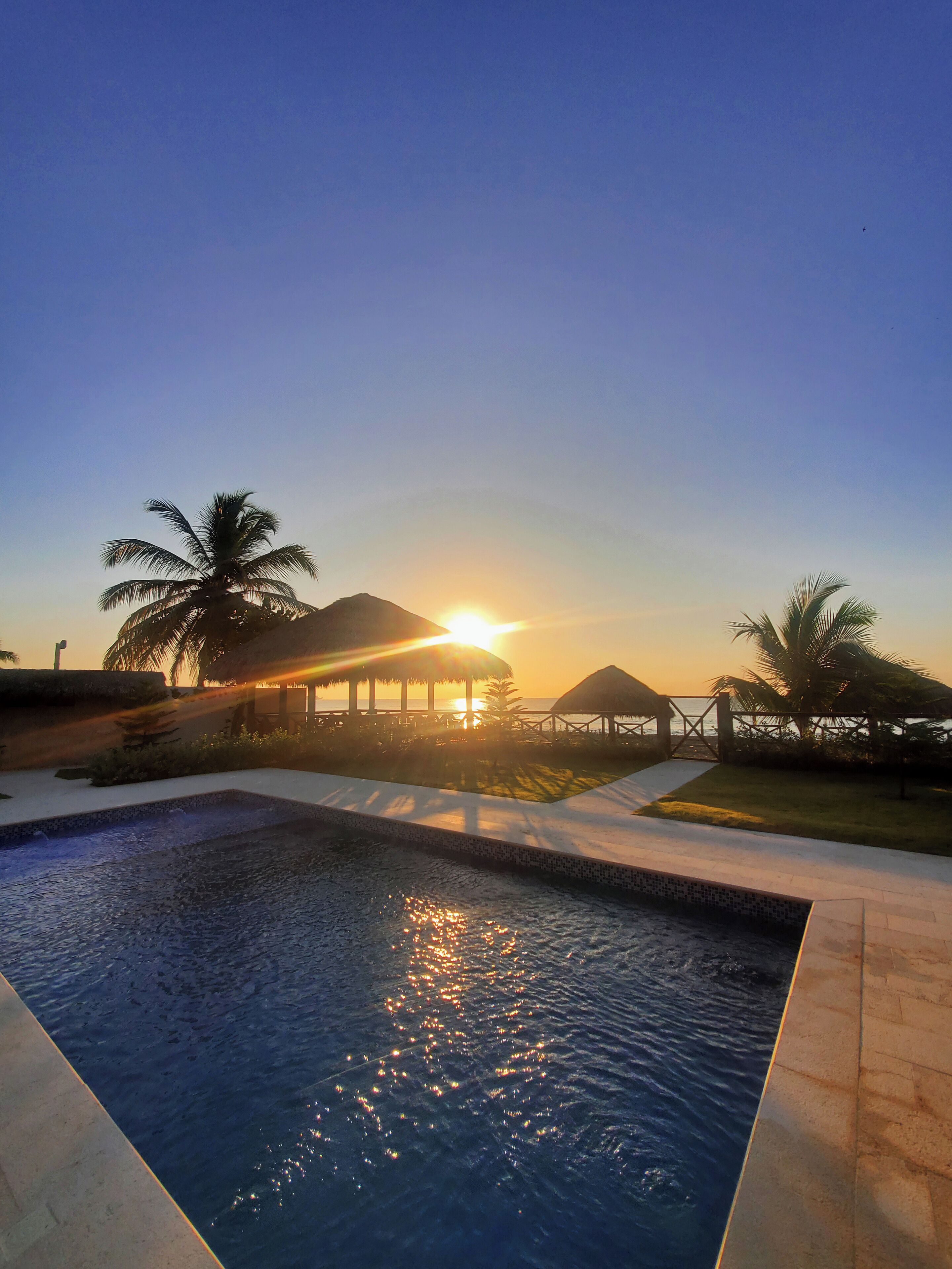A swimming pool with a sunset in the background, palm trees, and a thatched-roof structure.