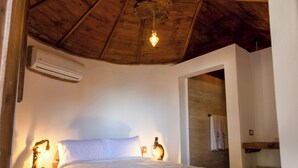 Un dormitorio con techo de madera, cama con ropa de cama blanca, mesita de noche con lámpara y aire acondicionado de pared.