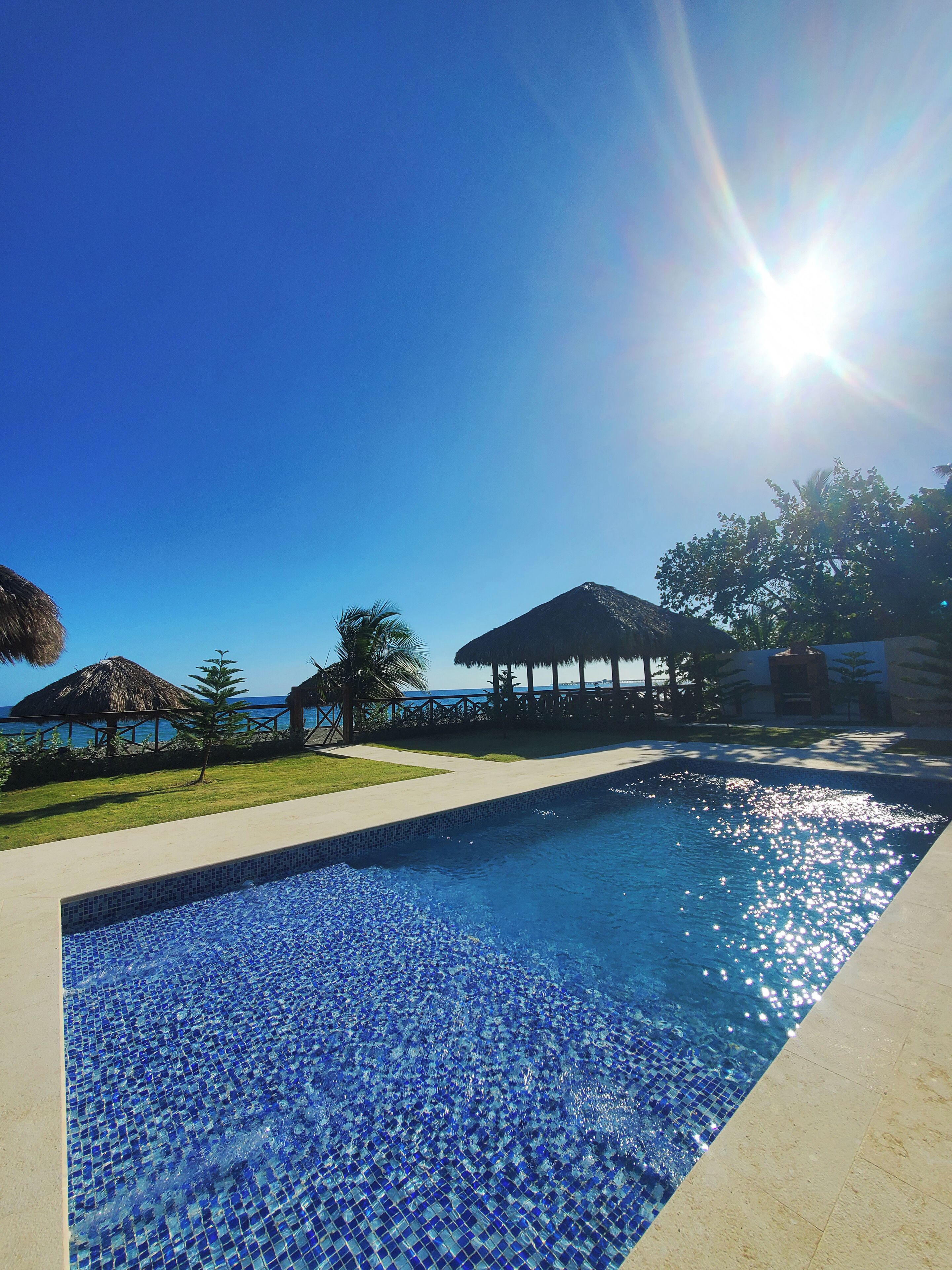 A swimming pool with clear blue water, surrounded by a concrete deck, palm trees, and a thatched-roof structure.