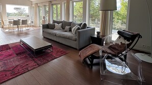 A modern living room with a grey sofa, a coffee table, and a clear chair. There are large windows providing natural light and a view of greenery outside.