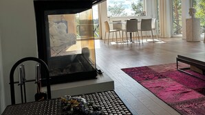 A modern living room with a fireplace, a dining area with a table and chairs, and a large window with a view of trees.