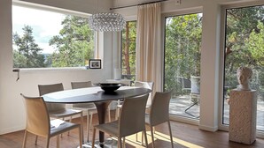 A modern dining room with a round table, six chairs, and a chandelier. Large windows offer a view of trees and a balcony.