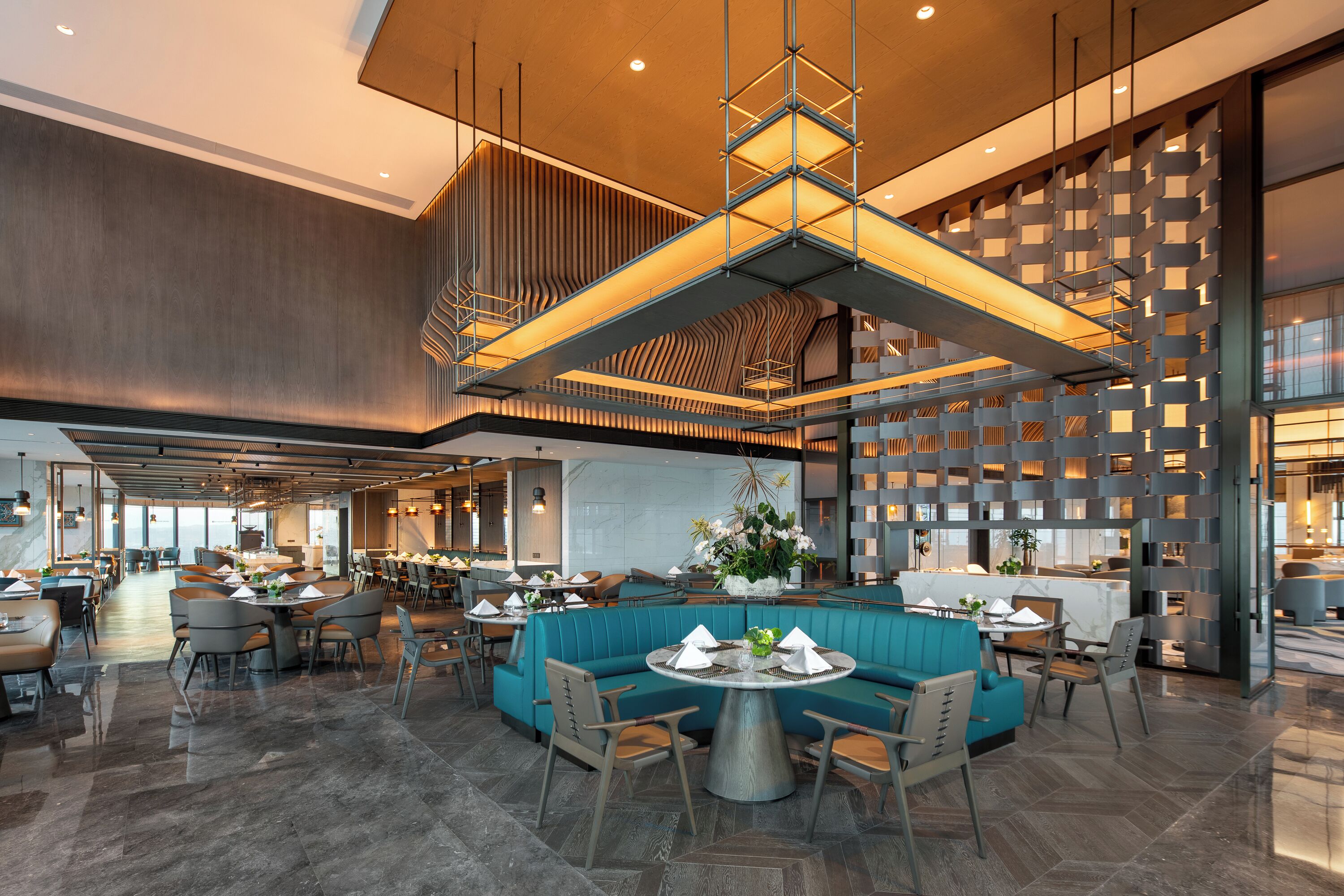 A spacious hotel lobby with a central seating area, multiple dining tables, and a modern ceiling design.