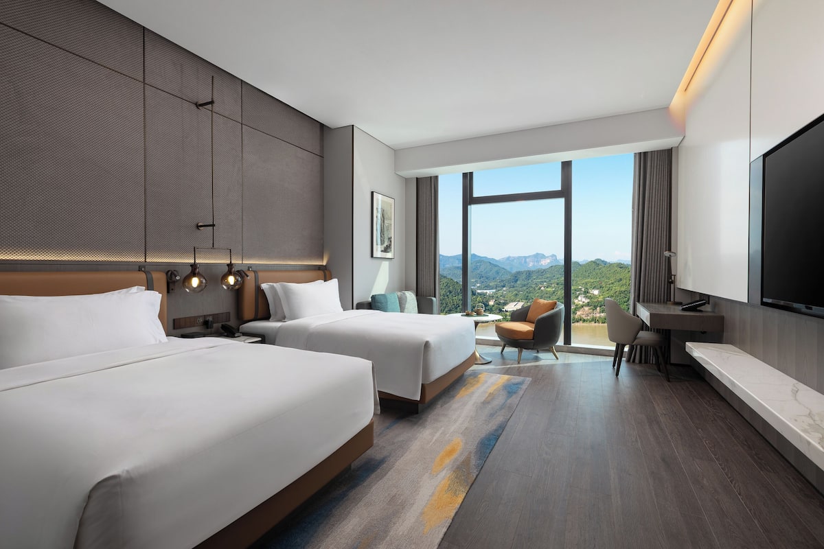 Habitación de hotel con dos camas, un escritorio, una silla, un televisor y un ventanal con vistas a la montaña.