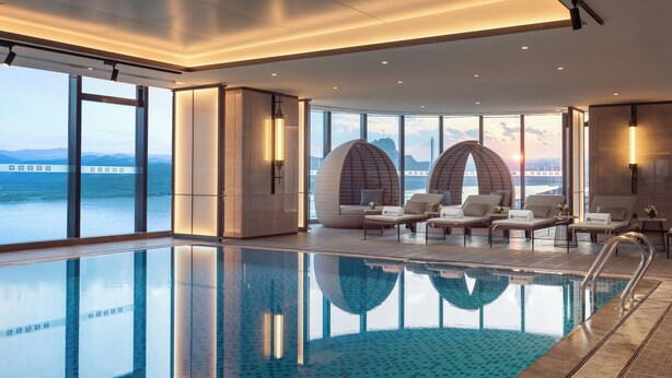 Una piscina interior moderna con paredes de vidrio transparente, sillones de descanso y vistas al mar.