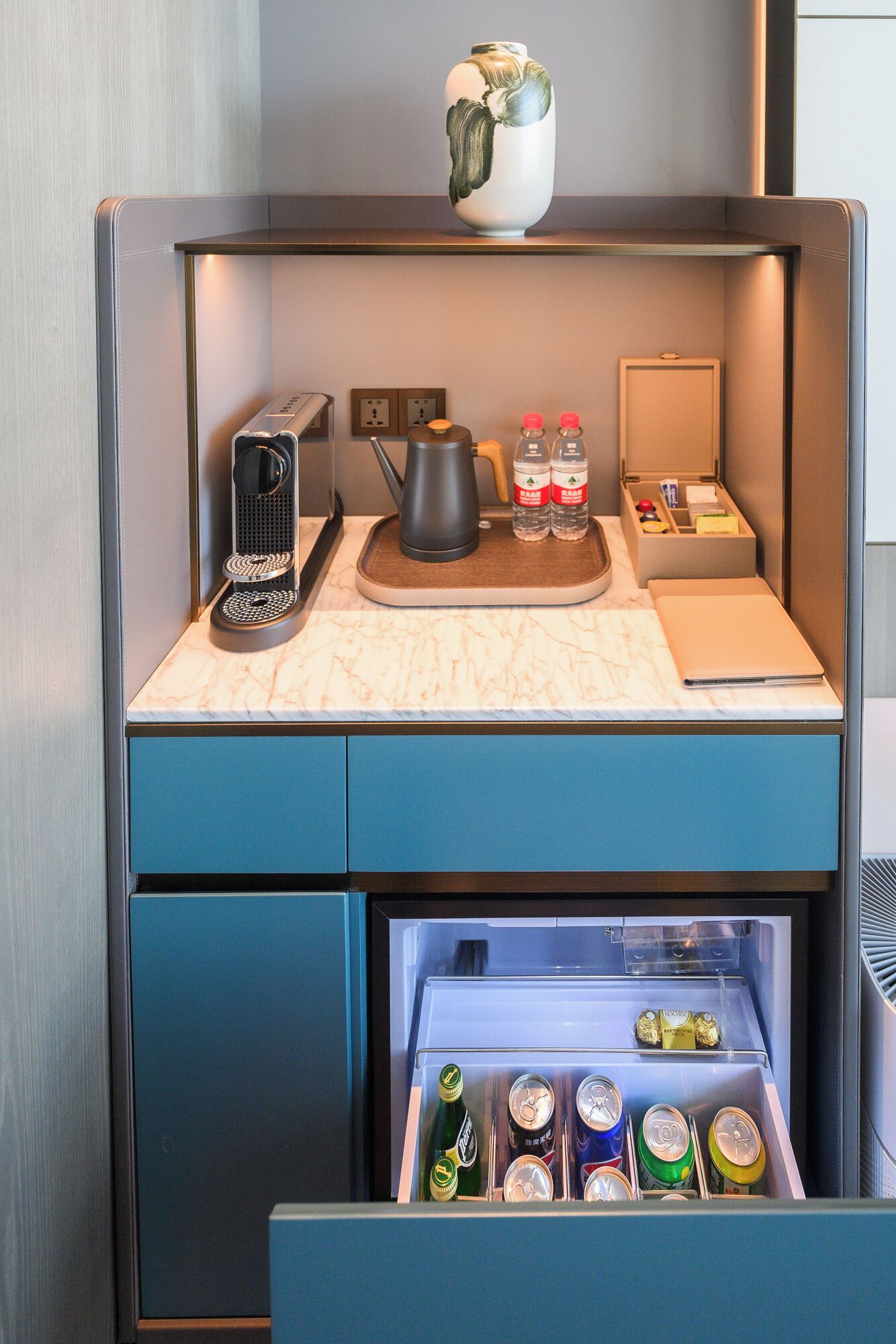 A compact kitchenette with a coffee maker, kettle, and a small refrigerator containing beverages.