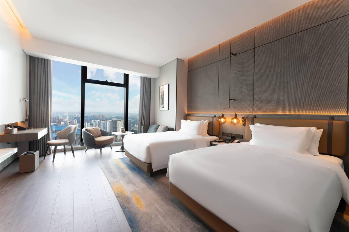 Habitación de hotel con dos camas, un escritorio, una silla y un ventanal con vistas a la ciudad.