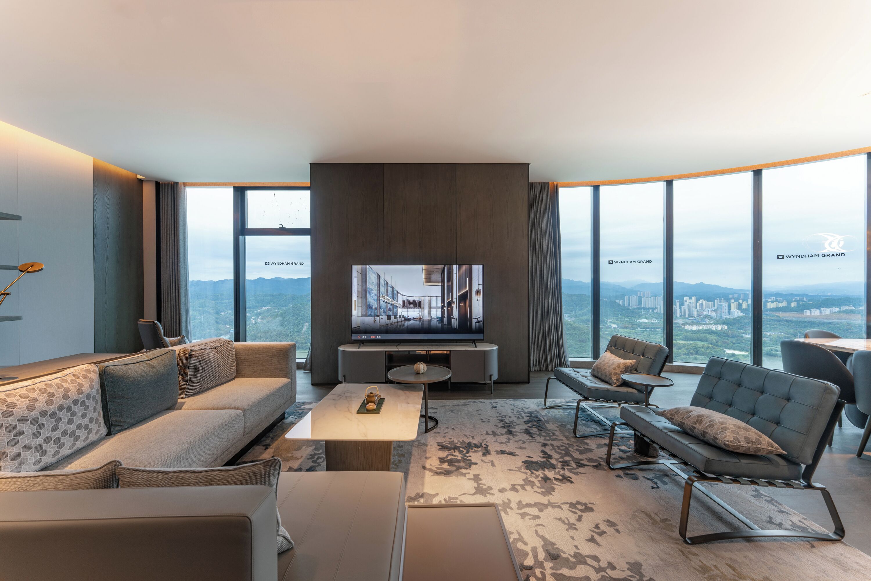 Una sala moderna con un televisor de pantalla plana grande, un sofá, una mesa de centro y vistas a un paisaje urbano a través de amplios ventanales.