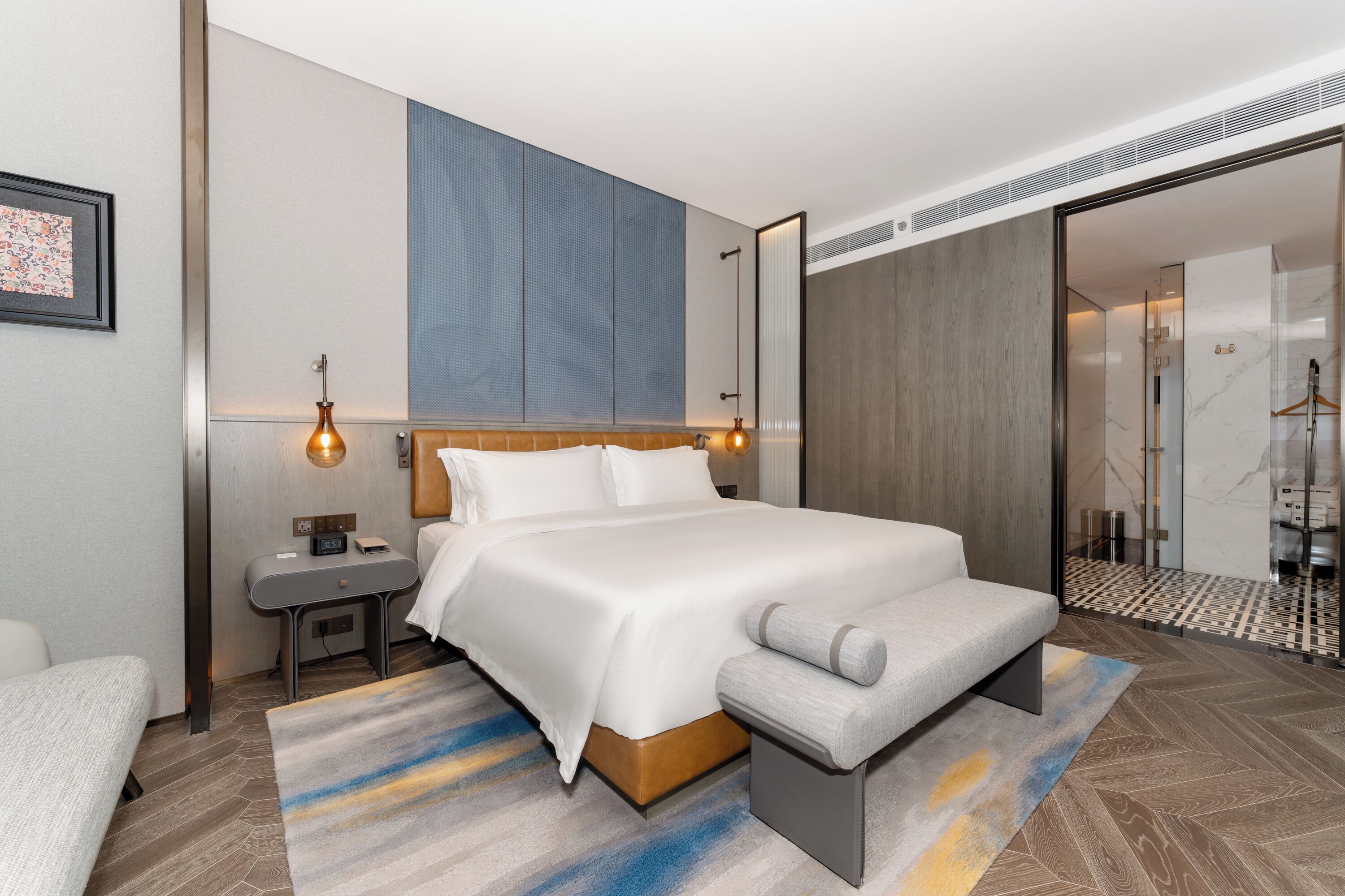 Una habitación de hotel moderna con una cama grande, mesitas de noche, un banco, zona de ducha y un espejo amplio.