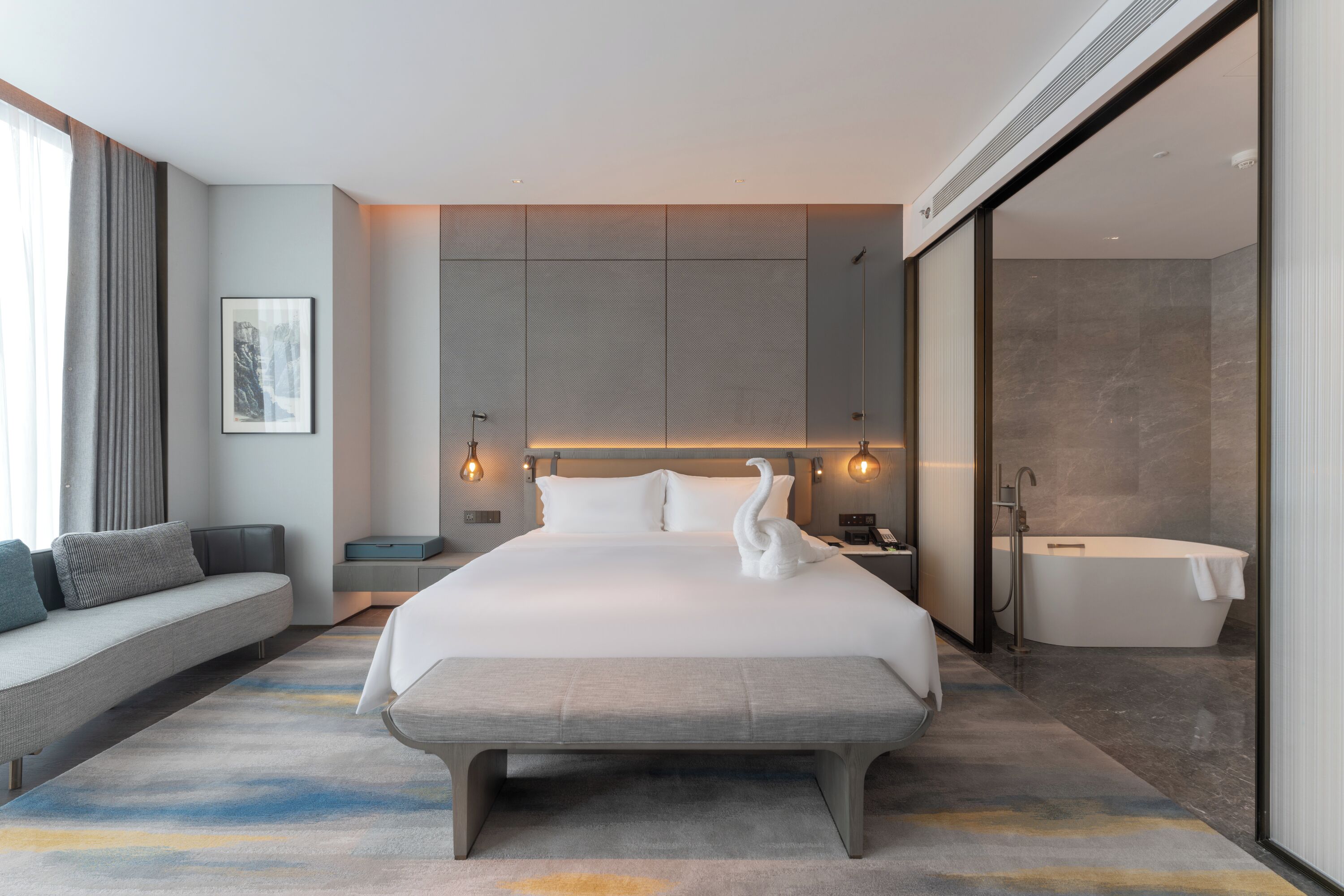 Una habitación de hotel moderna con una cama grande, un banco, un sofá, una bañera y un televisor montado en la pared.