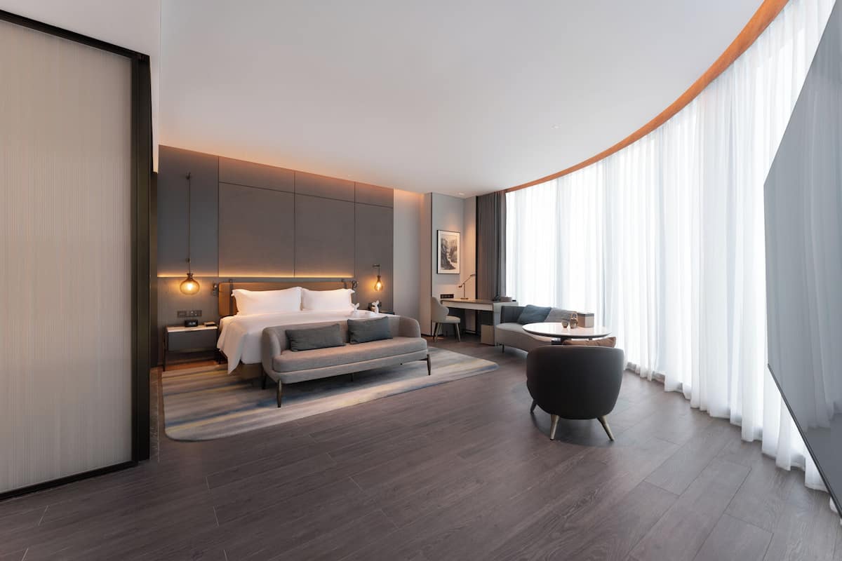 Una habitación de hotel moderna con cama, sofá, escritorio y un ventanal grande con cortinas.