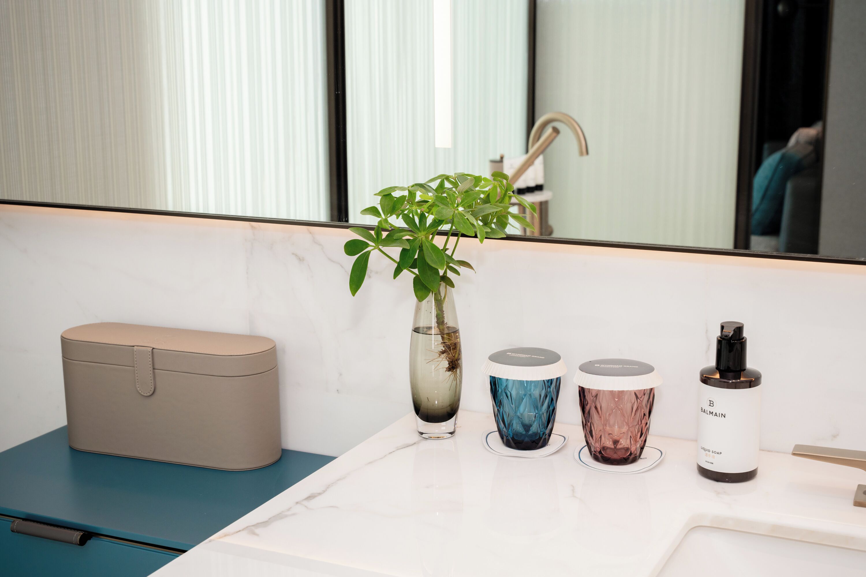 A bathroom vanity with a marble countertop, a beige toiletry bag, a potted plant, and a bottle of Balmain liquid soap.