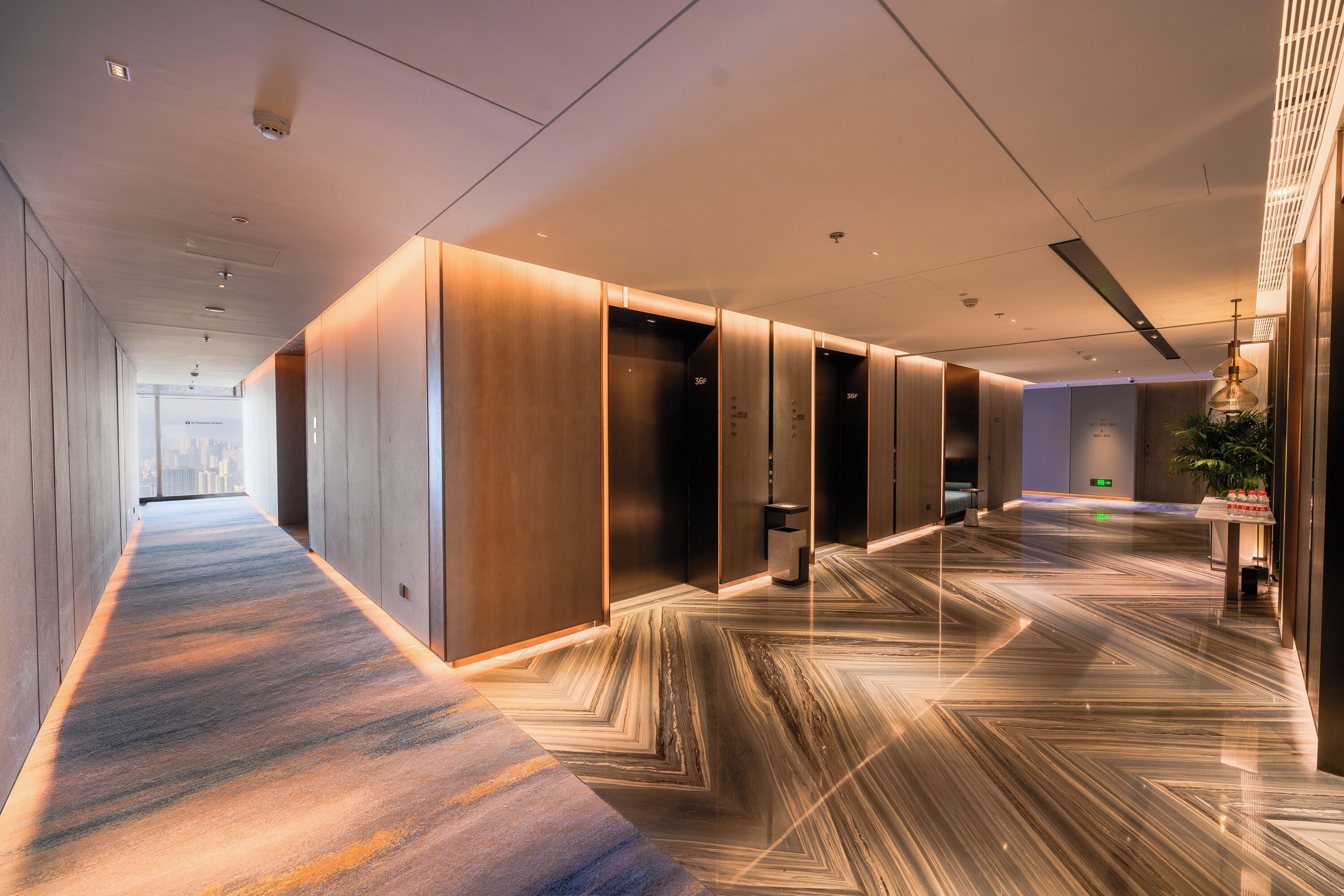Corredor moderno de hotel com piso estampado, iluminação embutida e vista para a paisagem urbana.