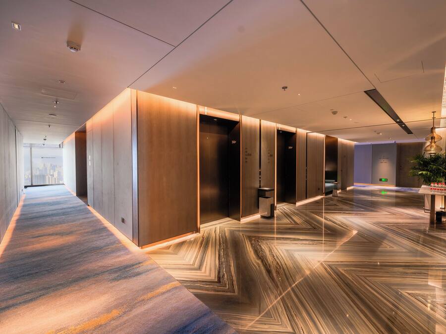 Corredor moderno de hotel com piso estampado, iluminação embutida e vista para a paisagem urbana.