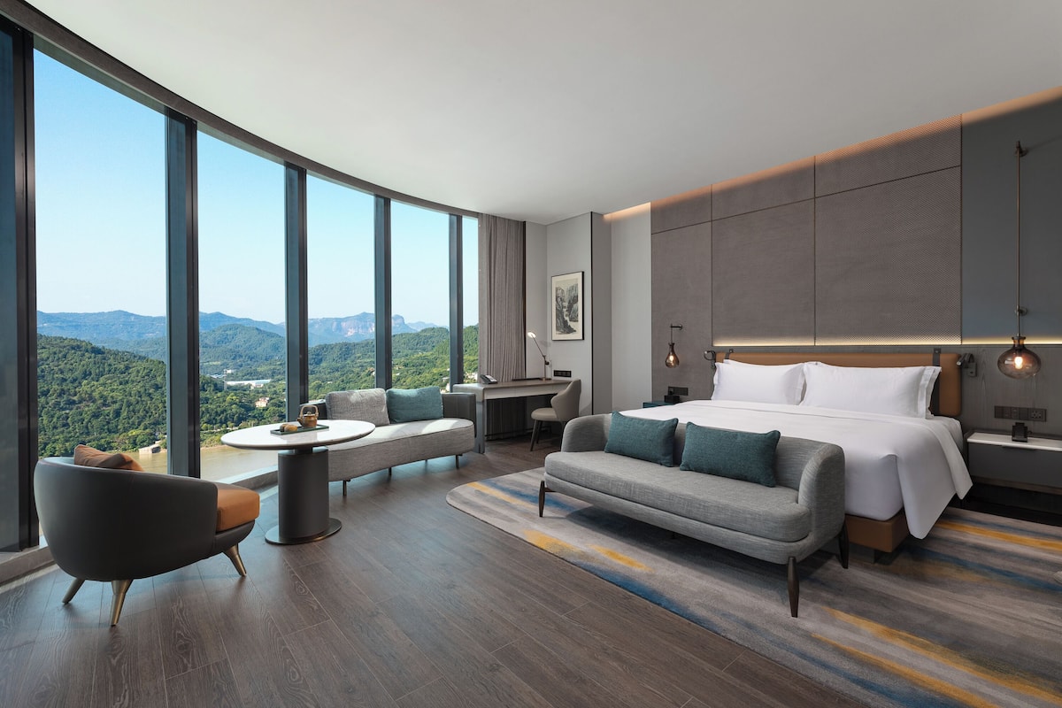 Habitación de hotel moderna con un ventanal grande, un sofá, una cama, un escritorio y vistas a las montañas.