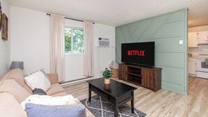 A living room with a TV displaying 'NETFLIX', a sofa, a coffee table, and a kitchen area in the background. - Walk to DT I King I Keyless Entry I Garage I W/D (Fargo)