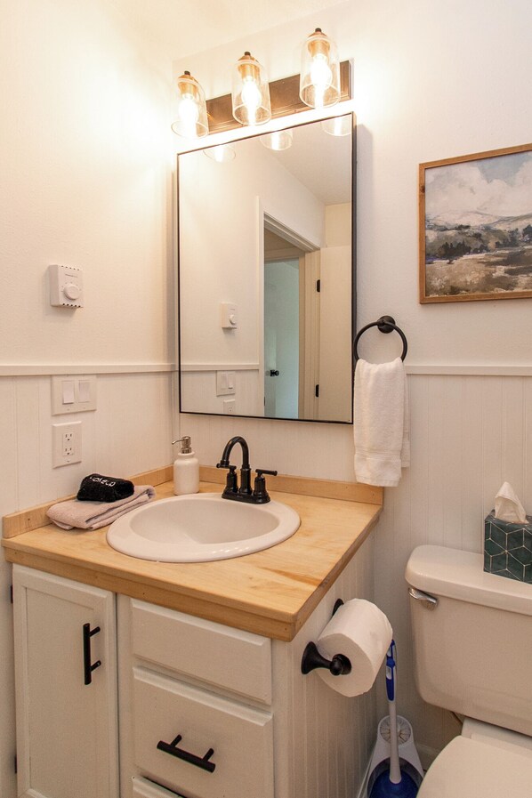 Ein Badezimmer mit einem Holzkosmetikschrank, einem Waschbecken, einem Spiegel, einem Handtuchhalter, einer Toilette und einem Bild an der Wand.