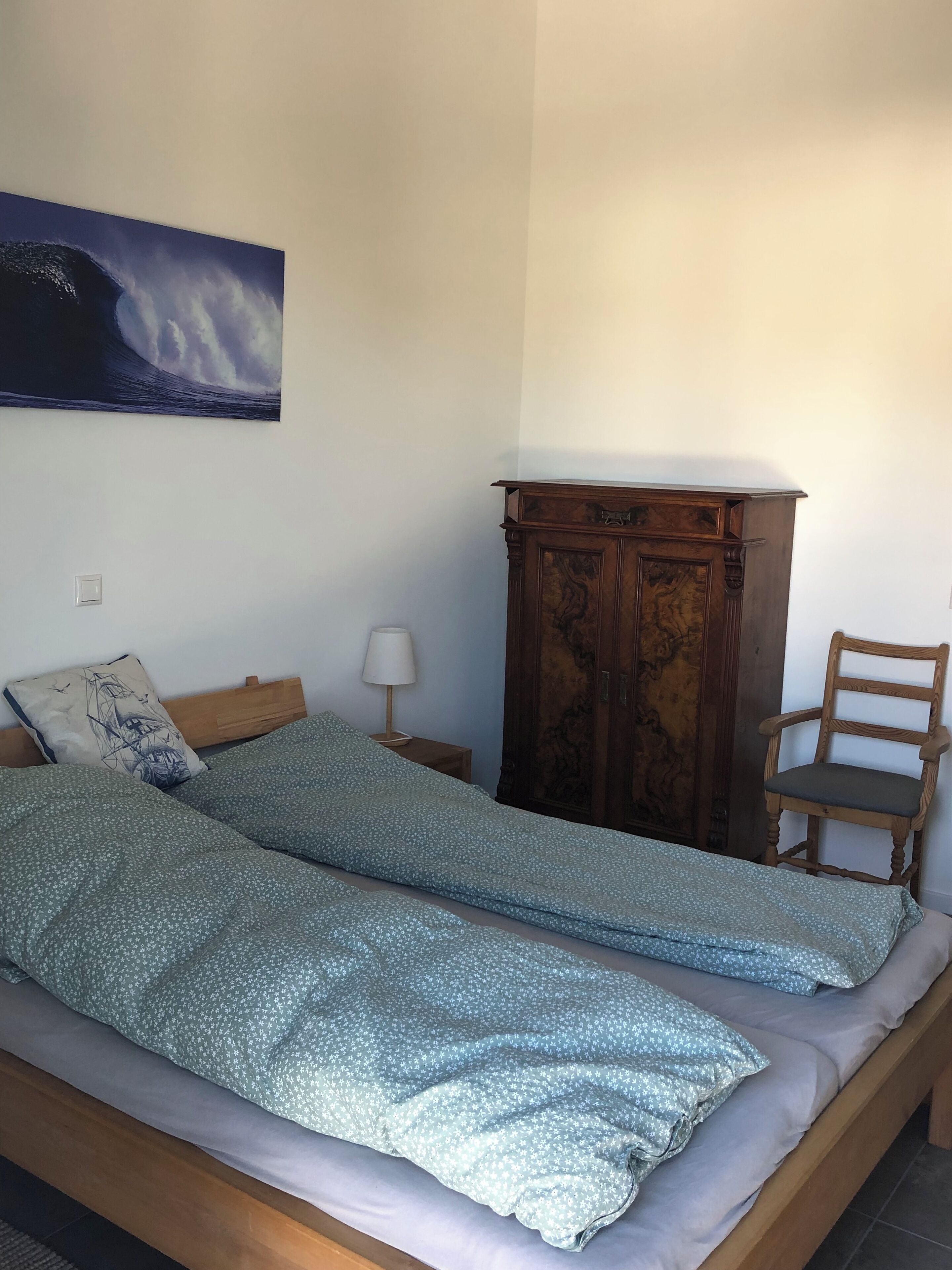A bedroom with a bed, a wooden wardrobe, a chair, a nightstand, a lamp, and a wall painting.