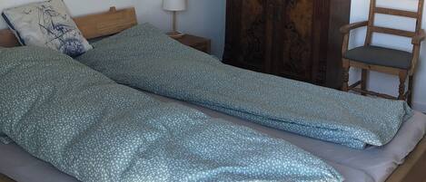 A bedroom with a bed, a wooden wardrobe, a chair, a nightstand, a lamp, and a wall painting.
