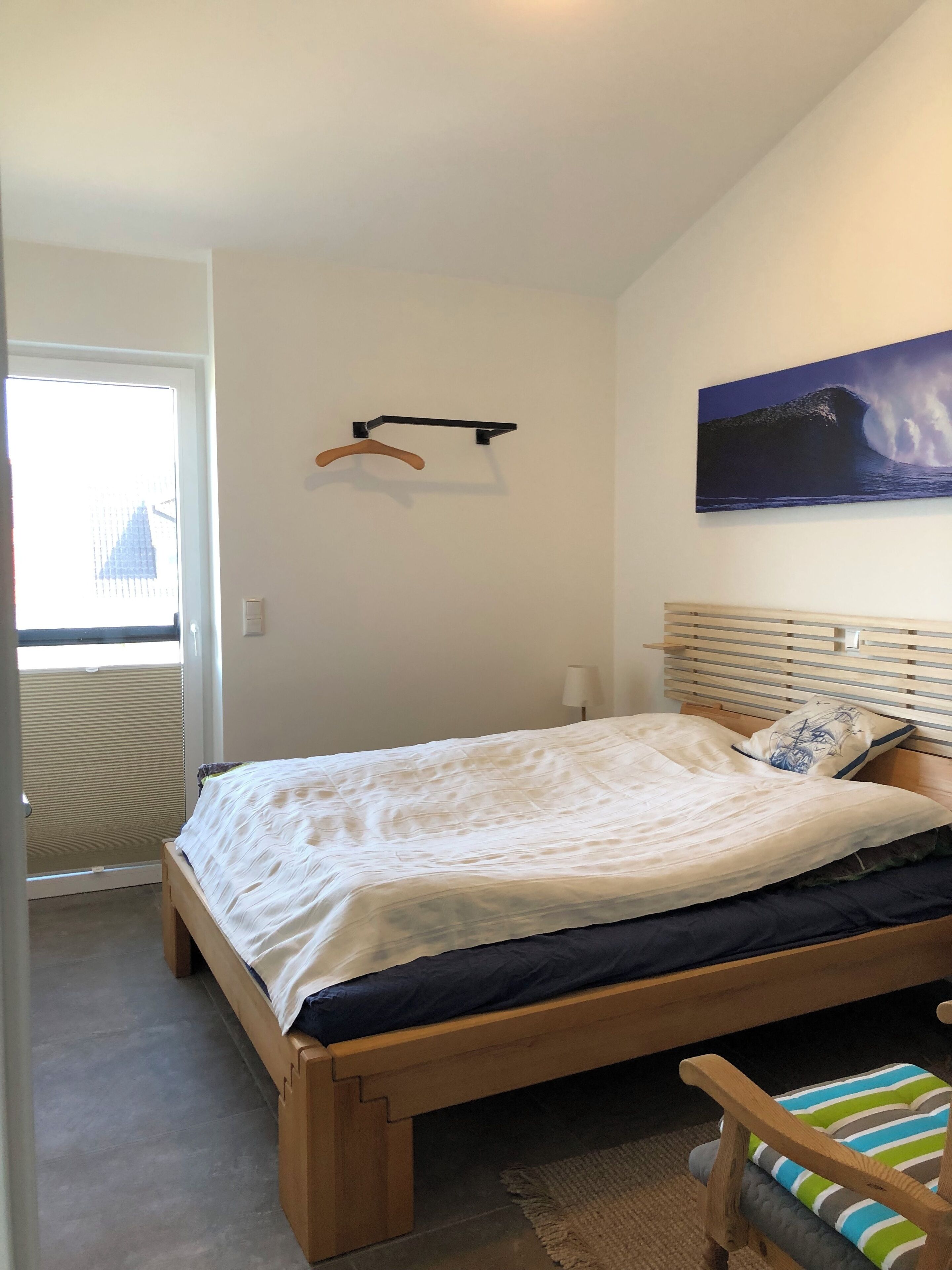 A bedroom with a bed, a wooden headboard, a wall-mounted coat rack, a painting, and a window with blinds.