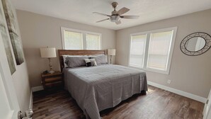 Une chambre à coucher comprenant un lit, des tables de chevet, un ventilateur de plafond et deux fenêtres équipées de stores.