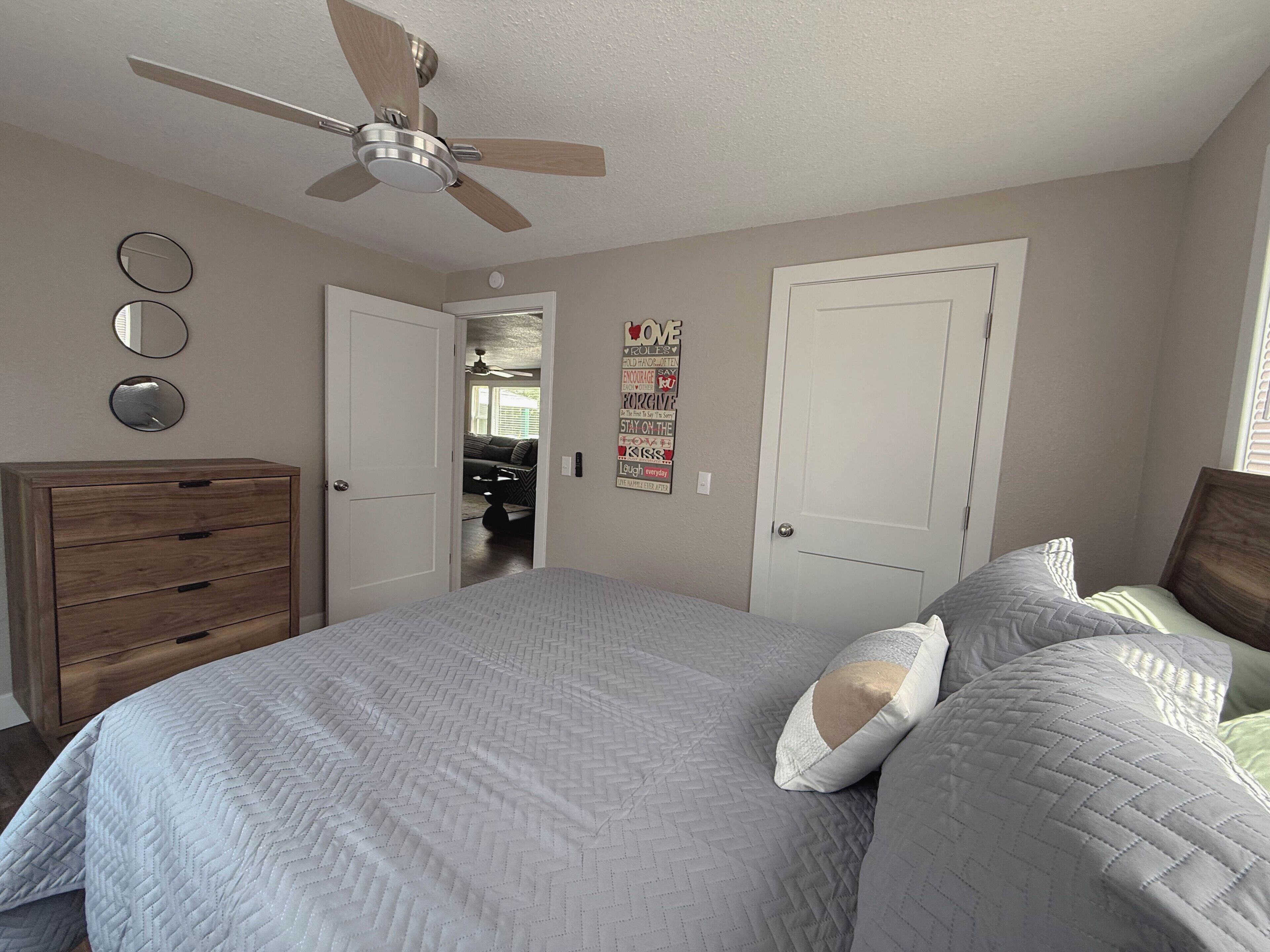 Ein Schlafzimmer mit einem Bett, einem Schminktisch, einem Deckenventilator und einer Tür zu einem anderen Raum.