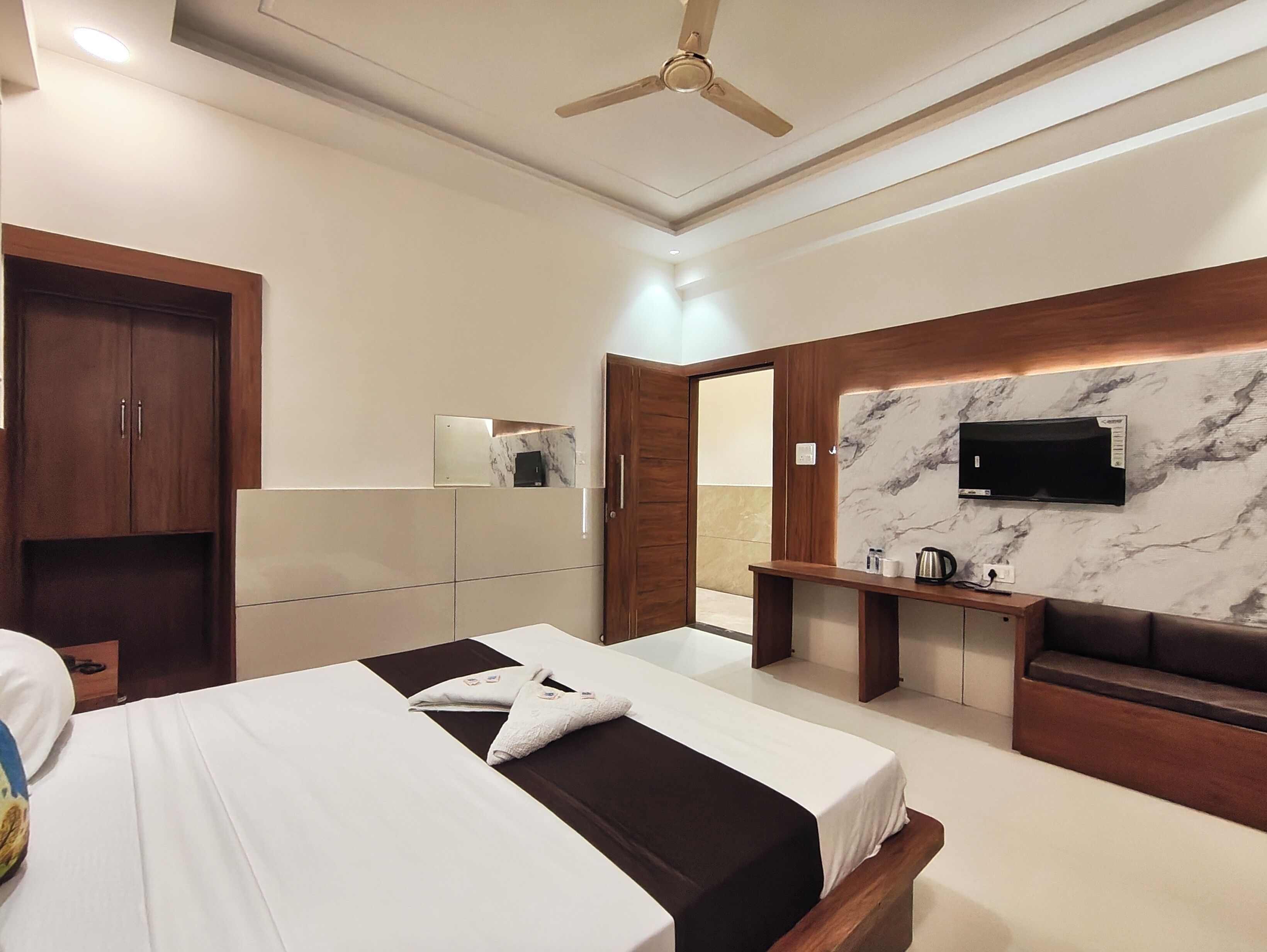 A modern hotel room with a bed, a wooden wardrobe, a TV mounted on a marble wall, and a ceiling fan.