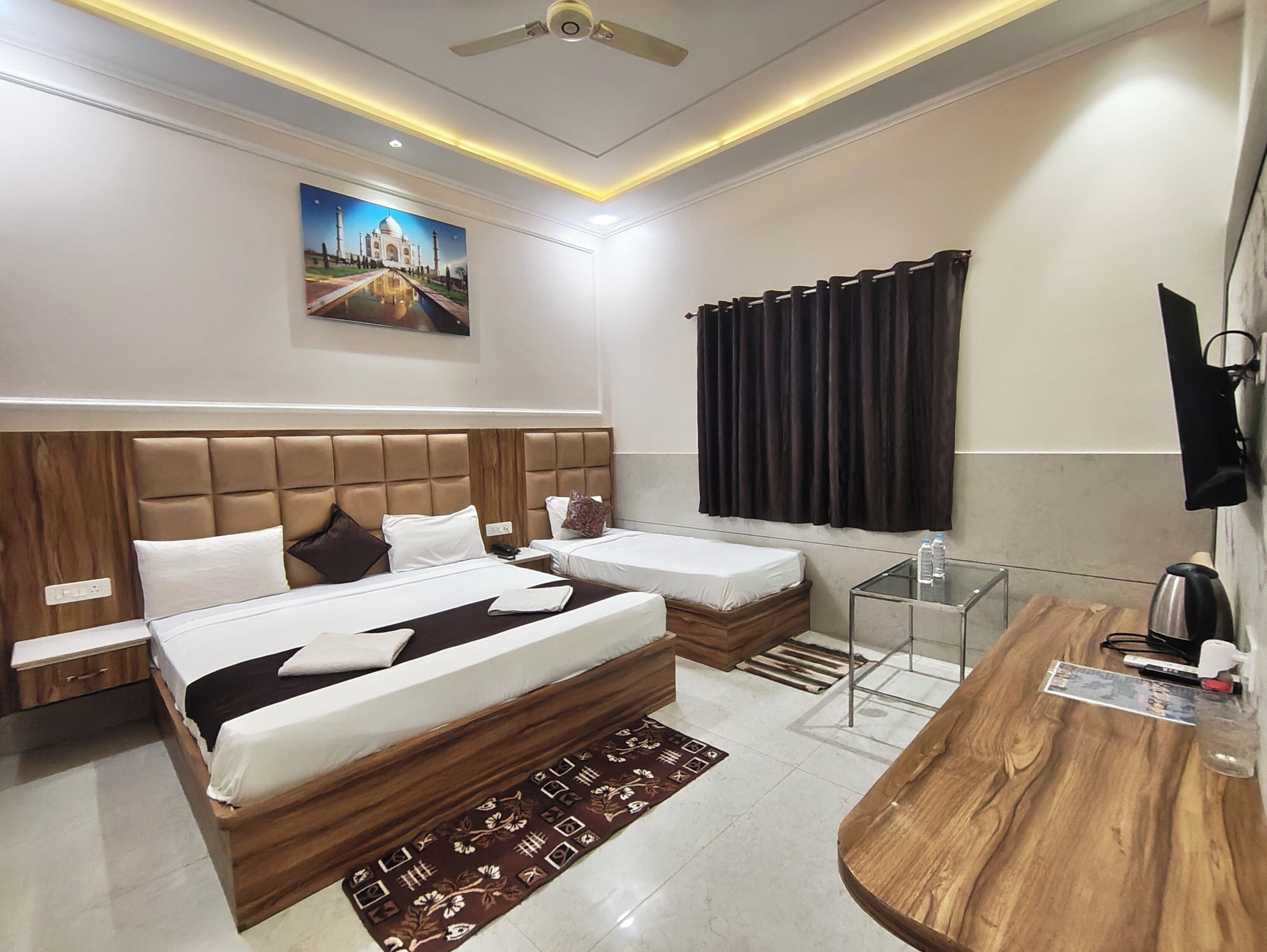 Una habitación de hotel moderna con dos camas, un cabecero de madera, un televisor de pantalla plana, un escritorio con un hervidor y un cuadro del Taj Mahal en la pared.