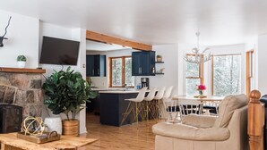 A modern kitchen with dark cabinets, a central island, and a dining area with white chairs. - 3 bedroom chalet on the river, indoor sauna, close to the ski village (Mont-Tremblant)